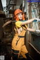 A woman in an orange hard hat and overalls holding a shovel.